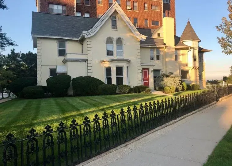 Cream-colored Victorian house with a red door, a black ornate fence, and well-kept lawn.