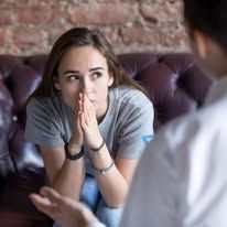 Woman with hands clasped, looking concerned, seated on a sofa, facing someone out of frame.