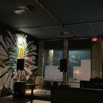 Interior of a dimly lit music venue with stage, microphone, and speakers. Background art on a wall.