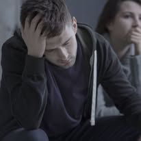 Man with head in hand, looking down, in distress. Woman in background, covering mouth.
