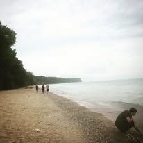 Beach scene with person crouching near water, others walking in distance. Overcast sky.