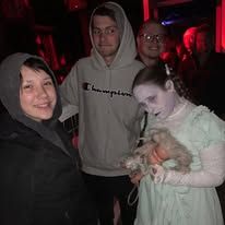 Three people pose: one in a Champion hoodie, one with pale makeup and costume, and another smiling with a hood.