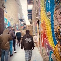 People walking in a narrow alley with colorful graffiti. One person in a brown hoodie, others are wearing coats.