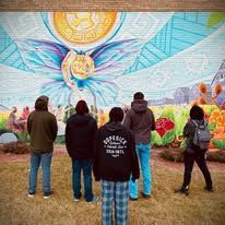 People looking at a colorful mural depicting a butterfly with a sun, in a grassy area with some buildings.