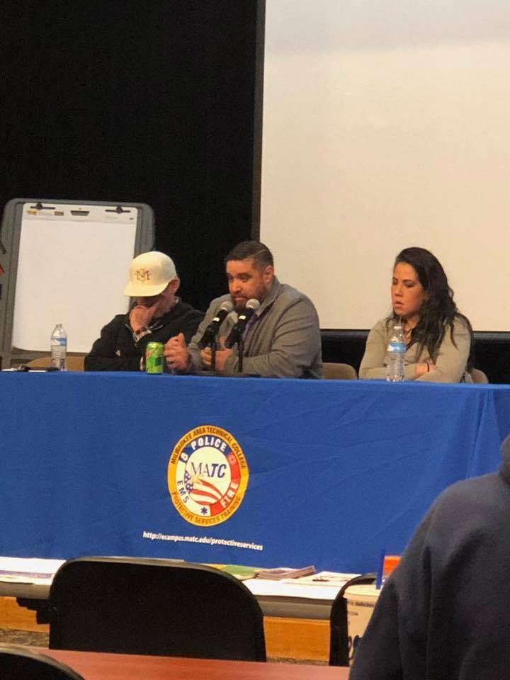 Three people seated at a table with microphones, speaking. Blue tablecloth with a logo.