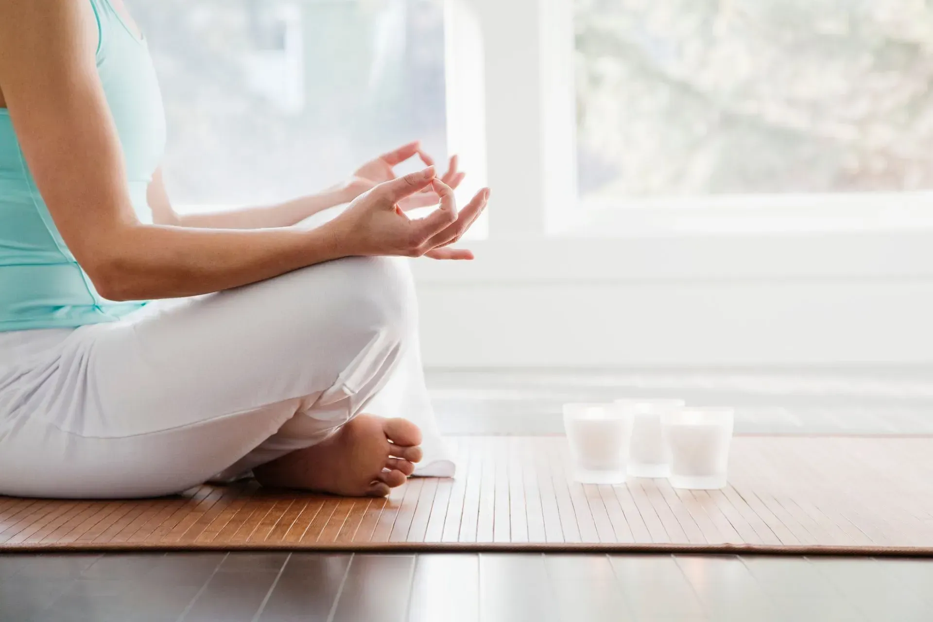 Person meditating indoors, legs crossed, hands in mudra, by a window, candles nearby.