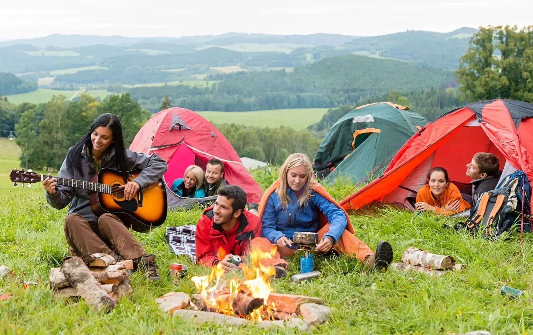 People camping in green field with tents, campfire, and playing guitar.