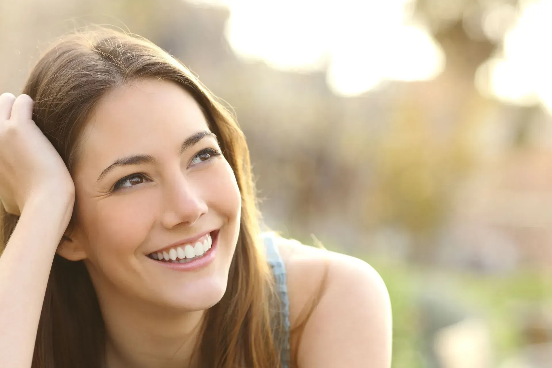 Woman smiling, looking up and to the right, hand on head, outdoor setting.