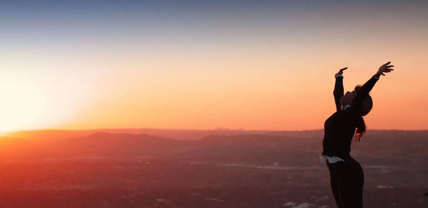 A person with arms raised in silhouette against a colorful sunset over a landscape.