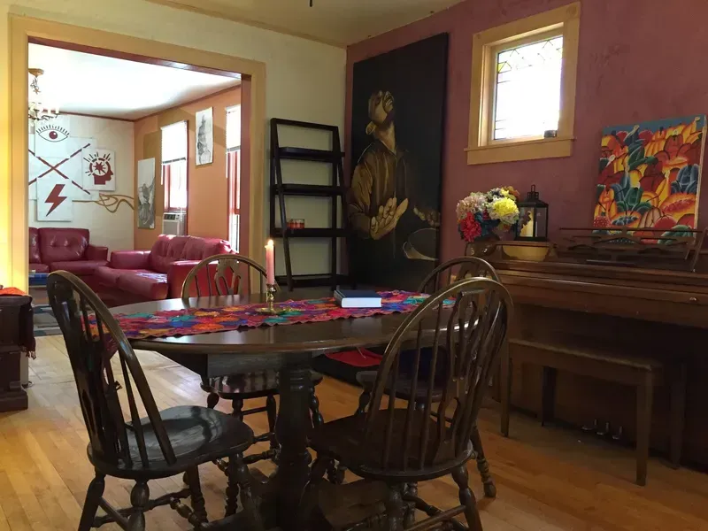 Dining room with wooden table and chairs, piano, painting, and doorway leading to living room with red furniture.
