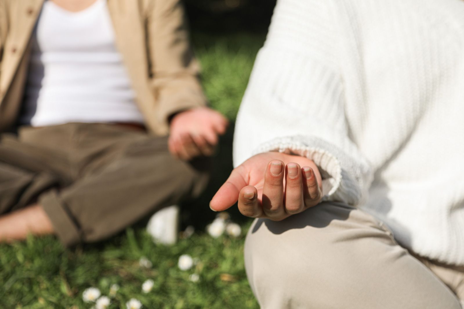 People meditating outdoors, one in a white sweater, hands in lap, focus on hand.