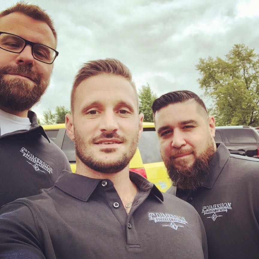 Three men in matching shirts smile outside near a yellow vehicle on an overcast day.