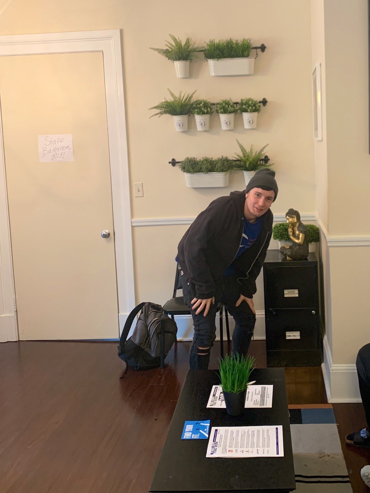 Man leans against table in waiting room, black jacket, ripped jeans, plants on wall.
