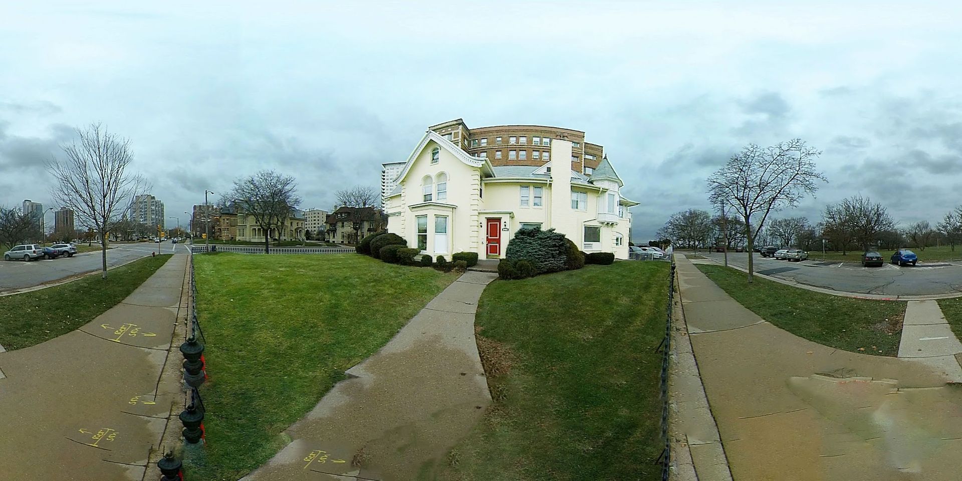 A white building with a damaged roof and a red door sits on a green lawn on a cloudy day.