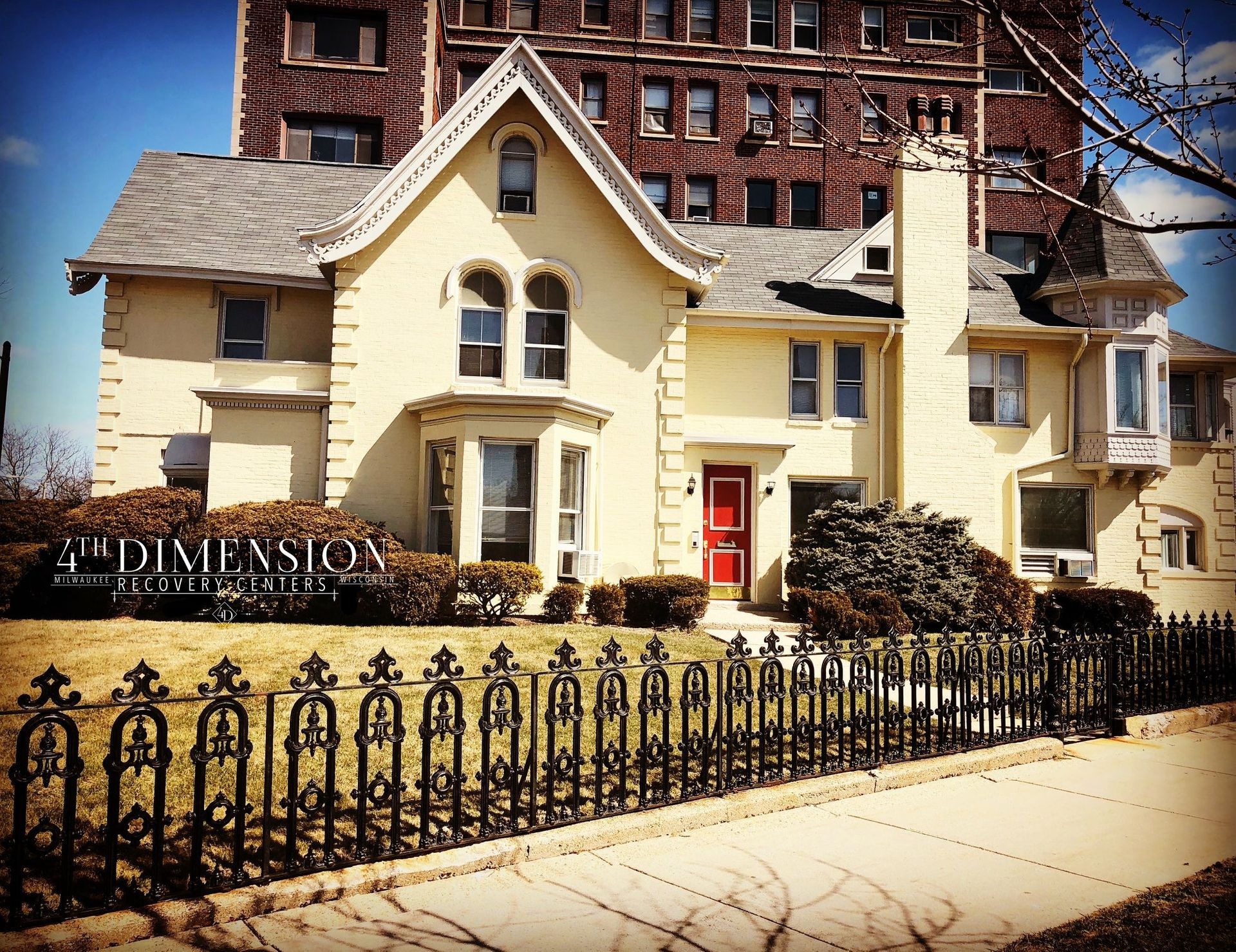 Yellow Victorian house with red door, black wrought-iron fence, and tall brick building in background.