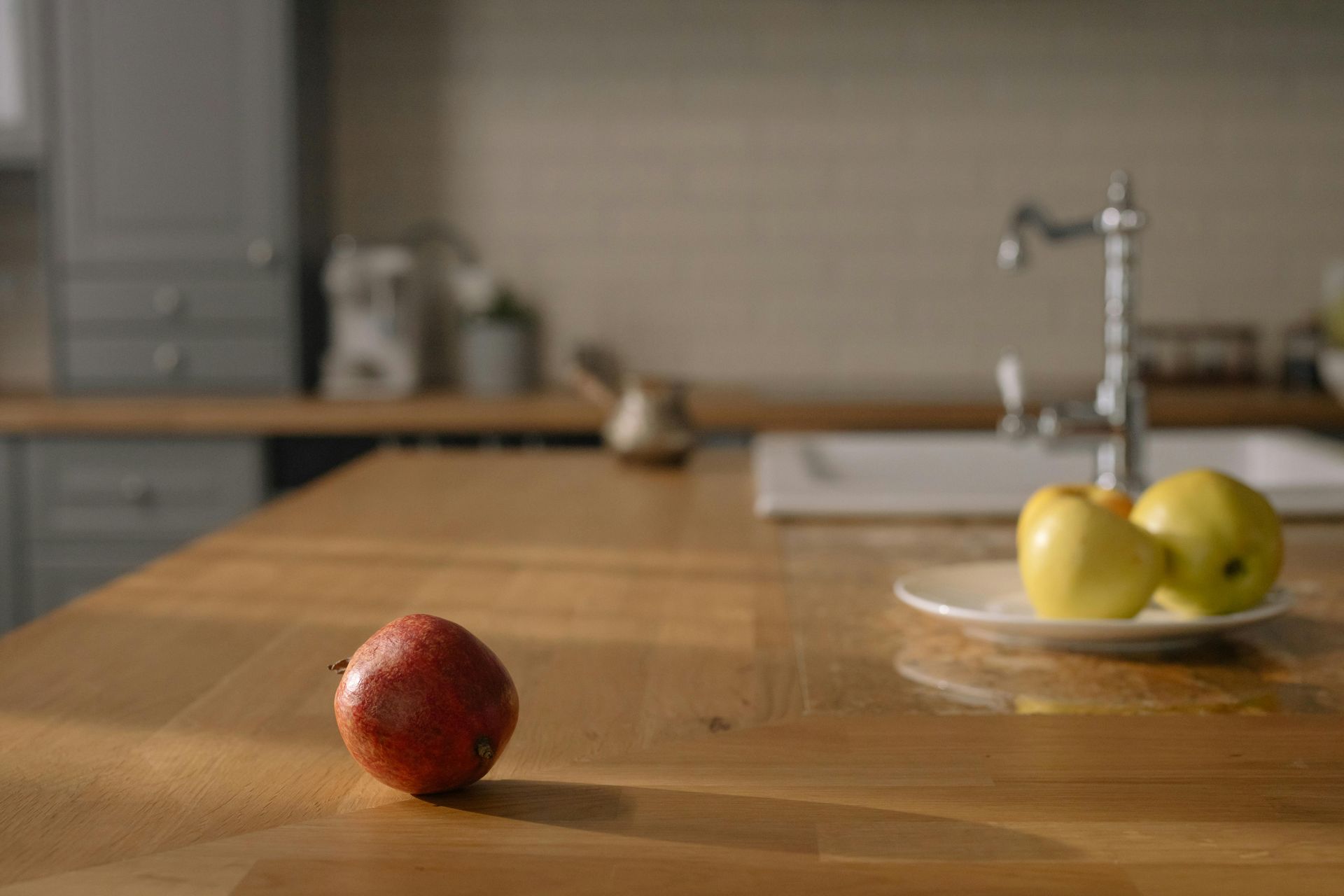 Red apple on a wooden kitchen counter with a bowl of green apples near a sink.