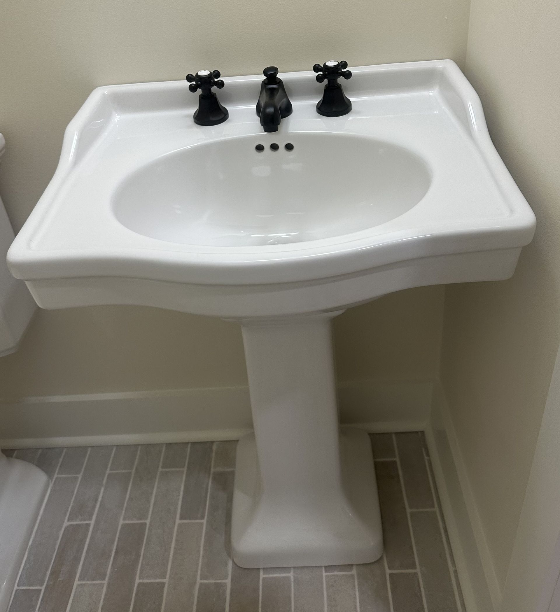 White pedestal sink with black faucets in a bathroom with gray tiled floor and beige walls.