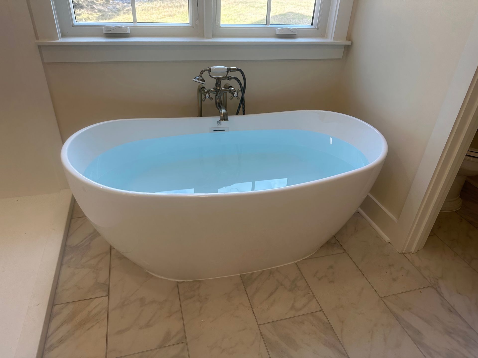 White oval bathtub filled with water by a window. Faucet visible. Marble tile flooring.