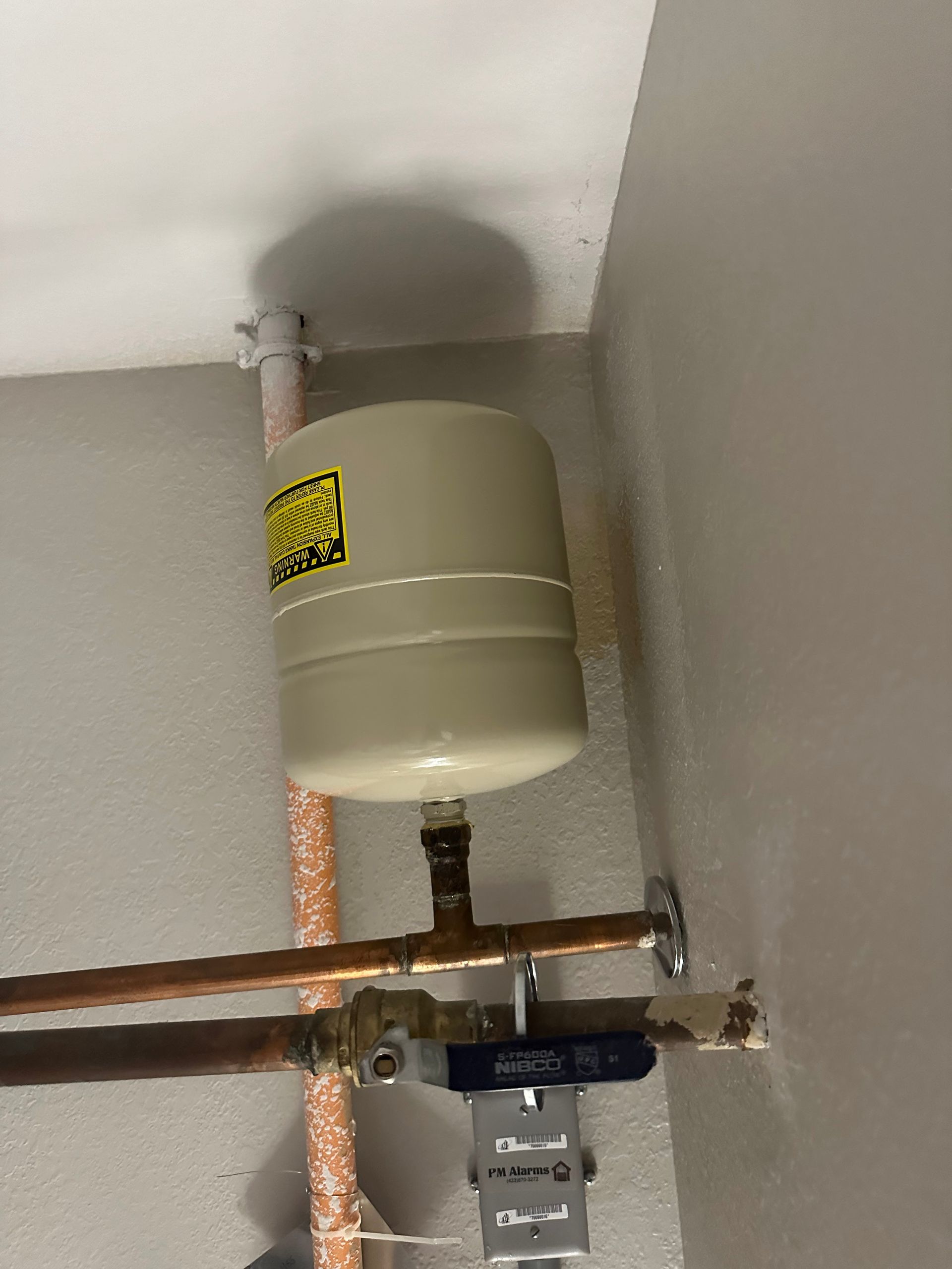 A cylindrical water tank with pipes in a utility room, mounted against a wall.