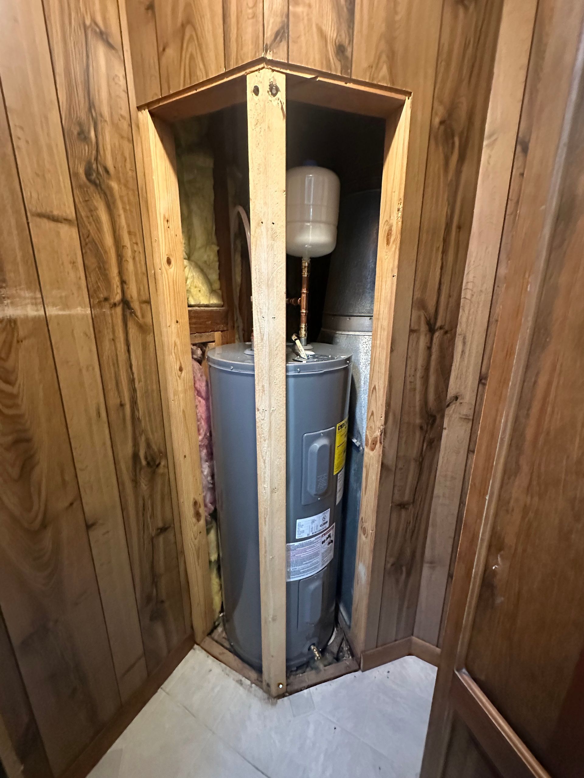 Water heater enclosed in wooden cabinet, placed in corner of room with wood paneling.