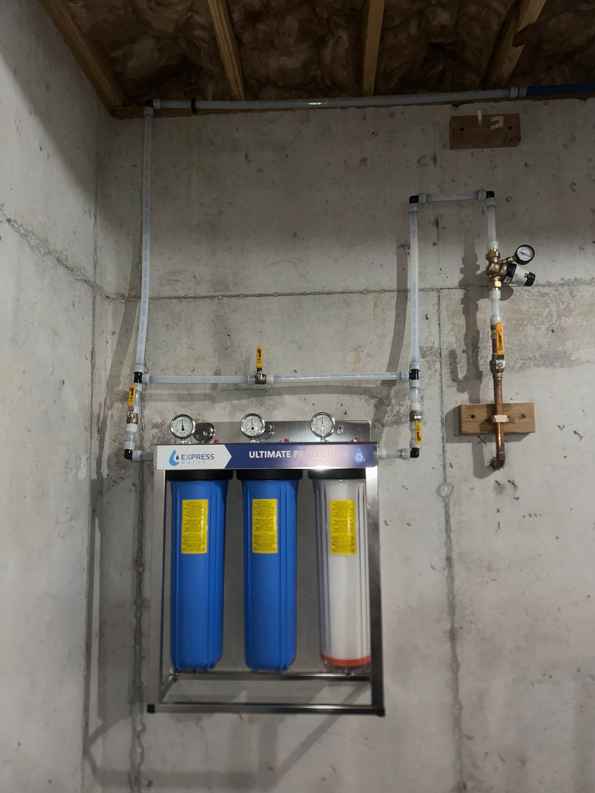 Water filtration system on a concrete wall, with blue and white filters and pressure gauges.