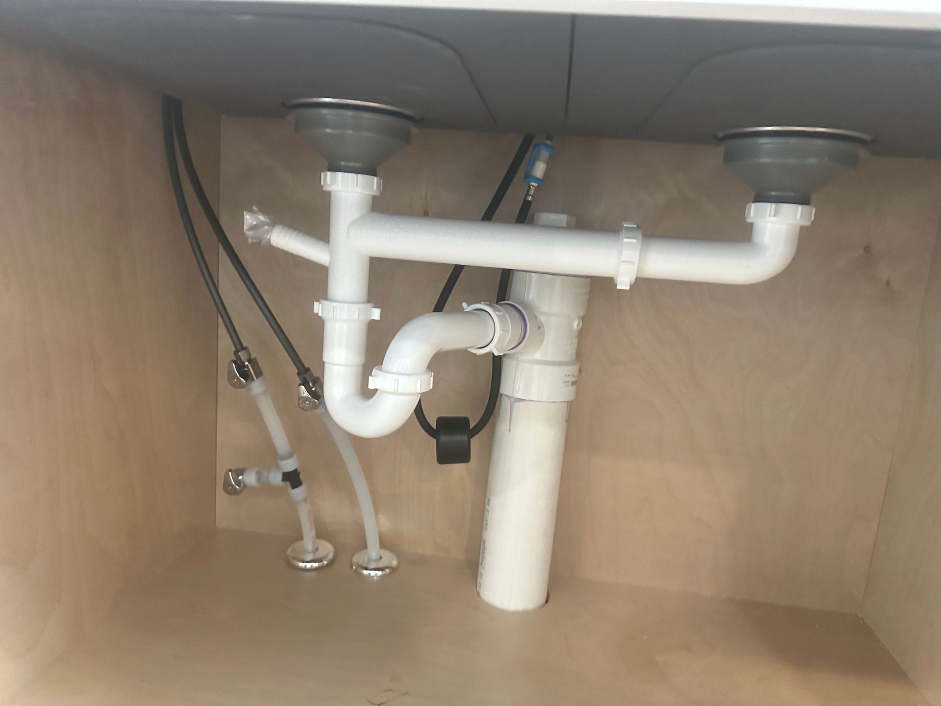 Undersink plumbing with white pipes, drain assemblies, and water supply lines within a wooden cabinet.