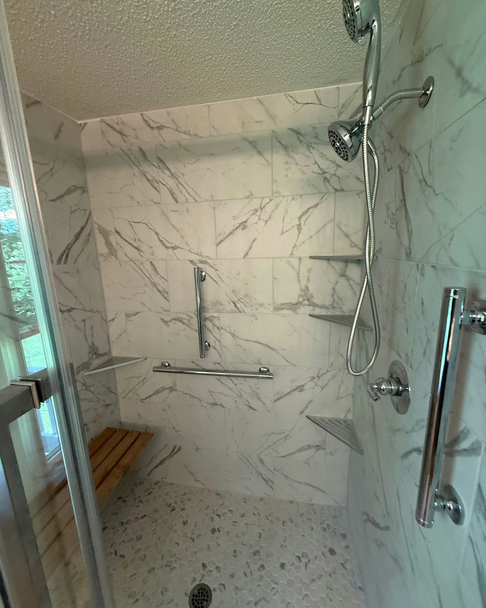 Accessible shower with marble tile, grab bars, a bench, and multiple shower heads.