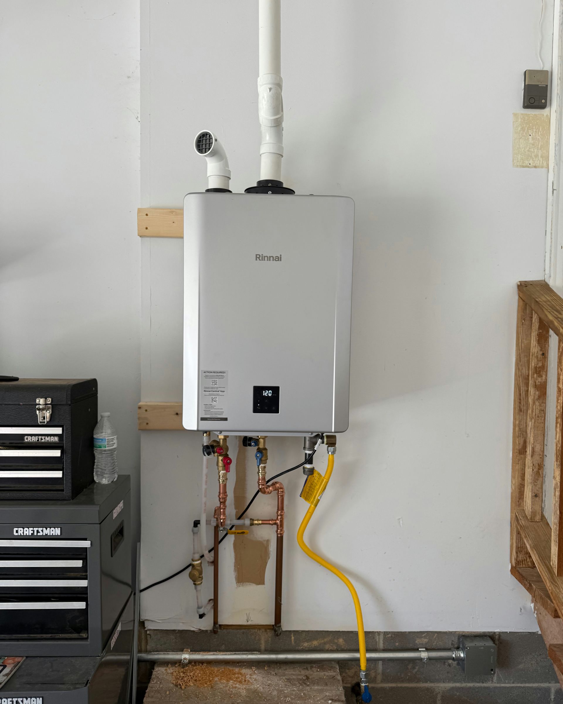 Rinnai tankless water heater mounted on a garage wall, with gas and water lines attached.