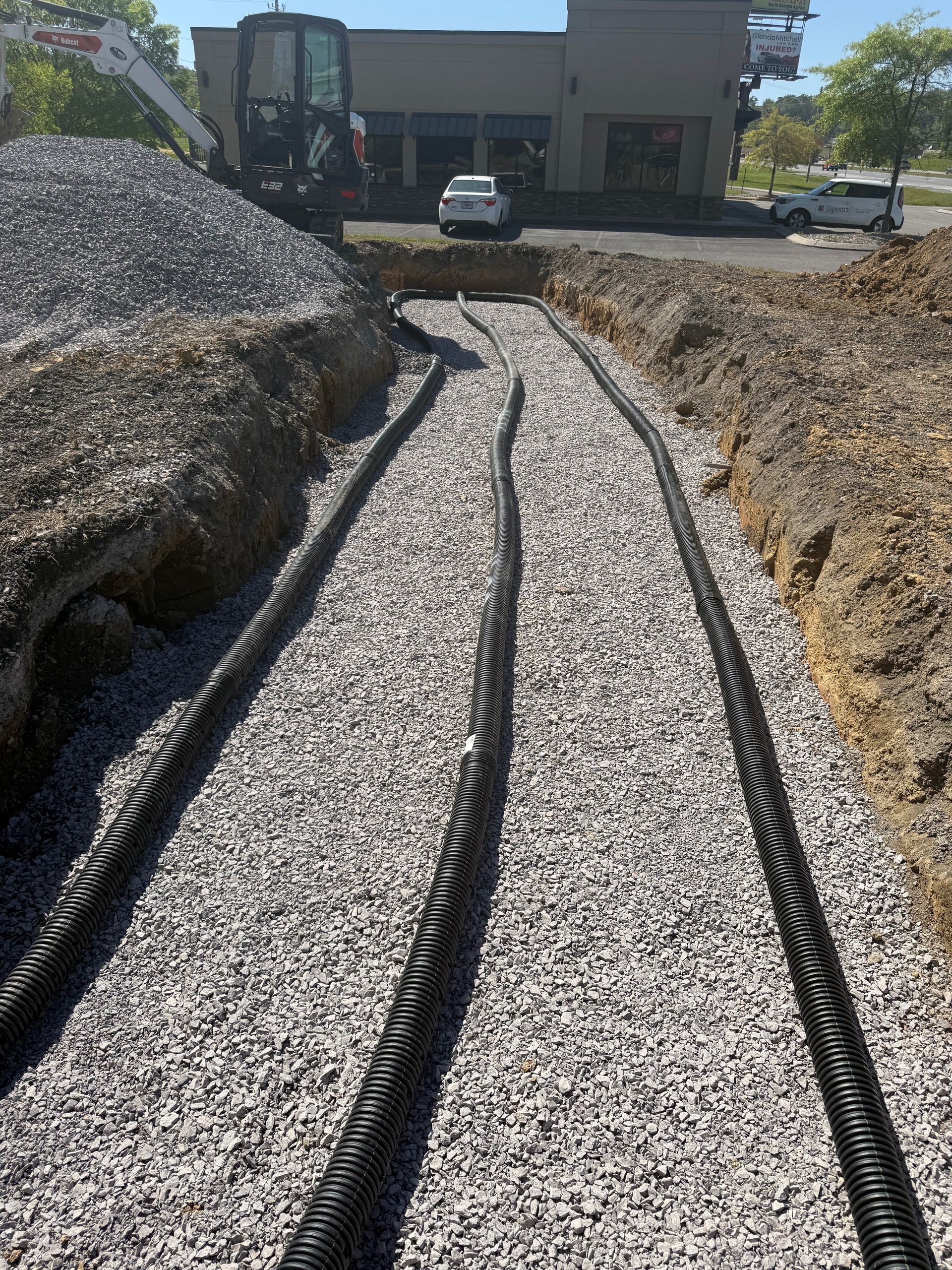 Trench with gravel and black pipes, likely for utility lines, near a commercial building under construction.