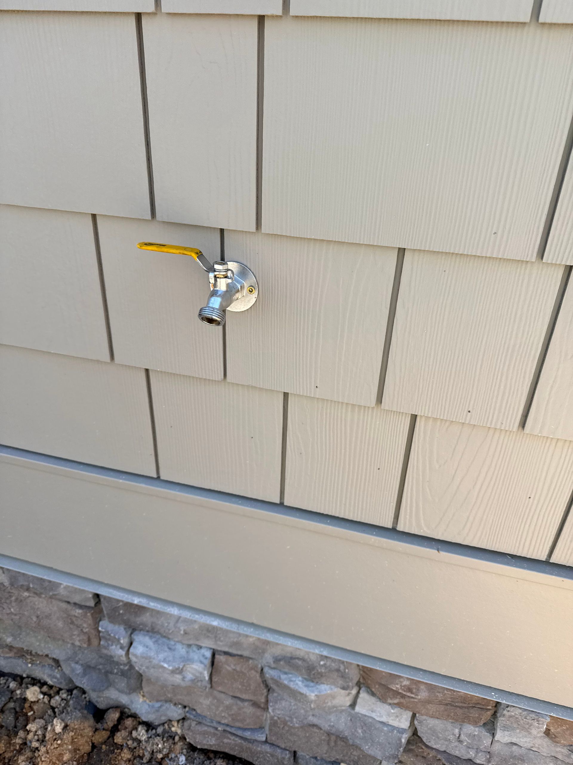 Exterior faucet on beige siding, mounted above a stone foundation.