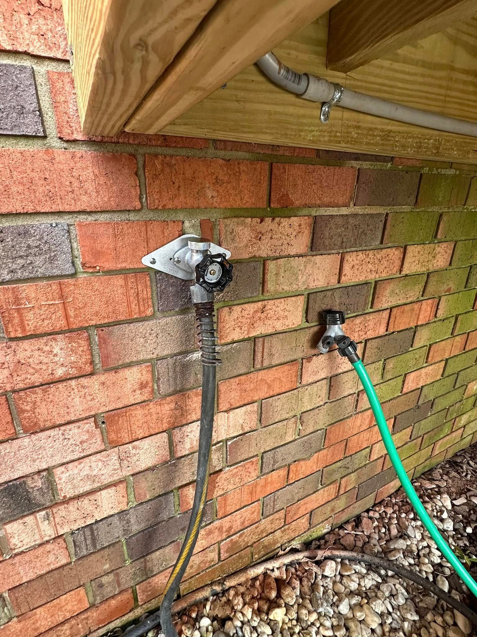 Brick wall with two outdoor water faucets, one with a black hose, one with a green hose.