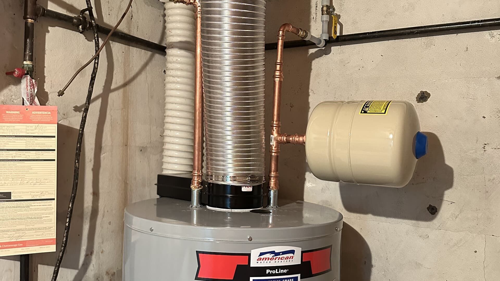 A water heater with copper piping, expansion tank, and insulation against a concrete wall.