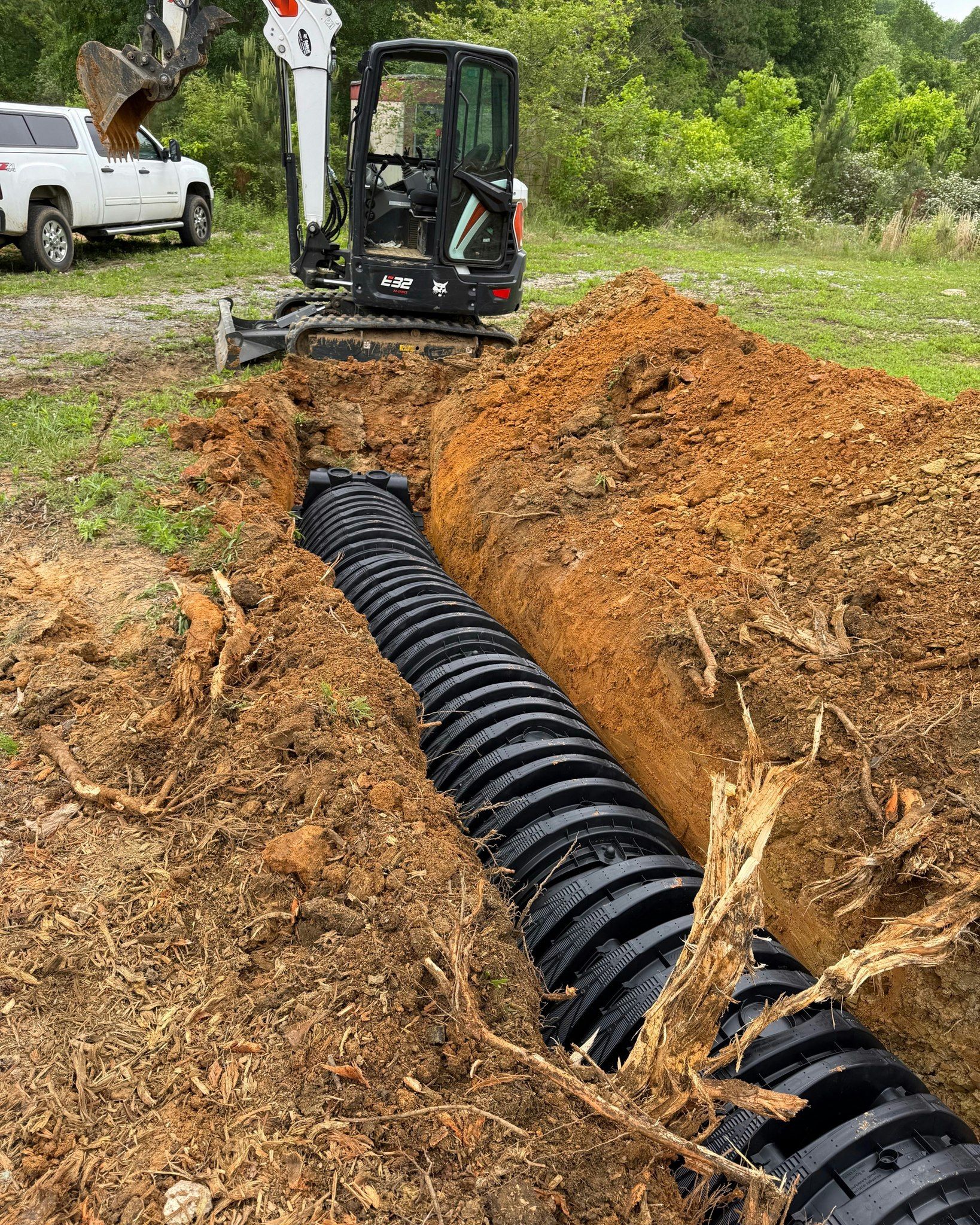 Black corrugated pipe in a trench, being installed by an excavator. A white pickup truck sits in the background.