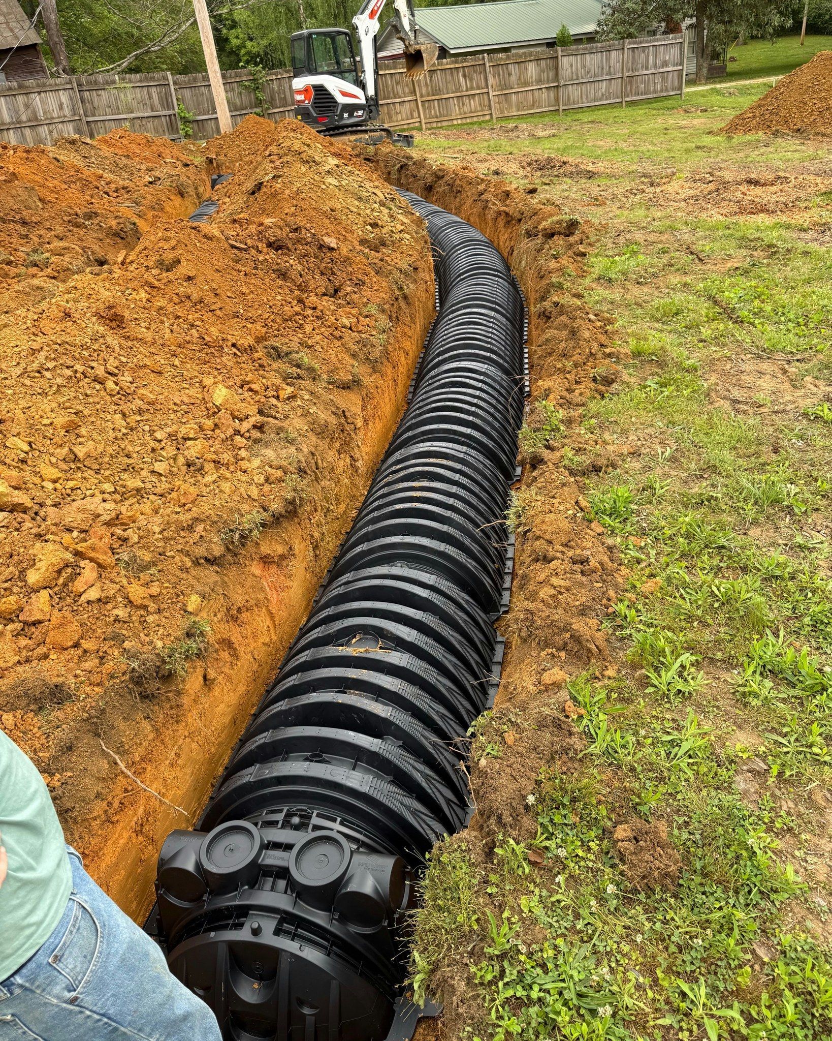 Black drainage pipe installed in a trench in a yard; a mini excavator is in the background.