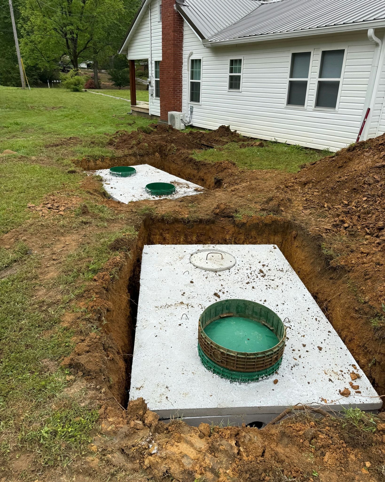 Excavated ground with septic tanks, grass, and a white house in the background.