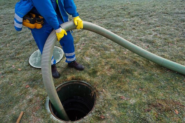 Person in blue overalls and yellow gloves emptying septic tank with large hose.
