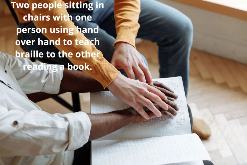 Two people sitting with one person teaching braille using hand over hand.