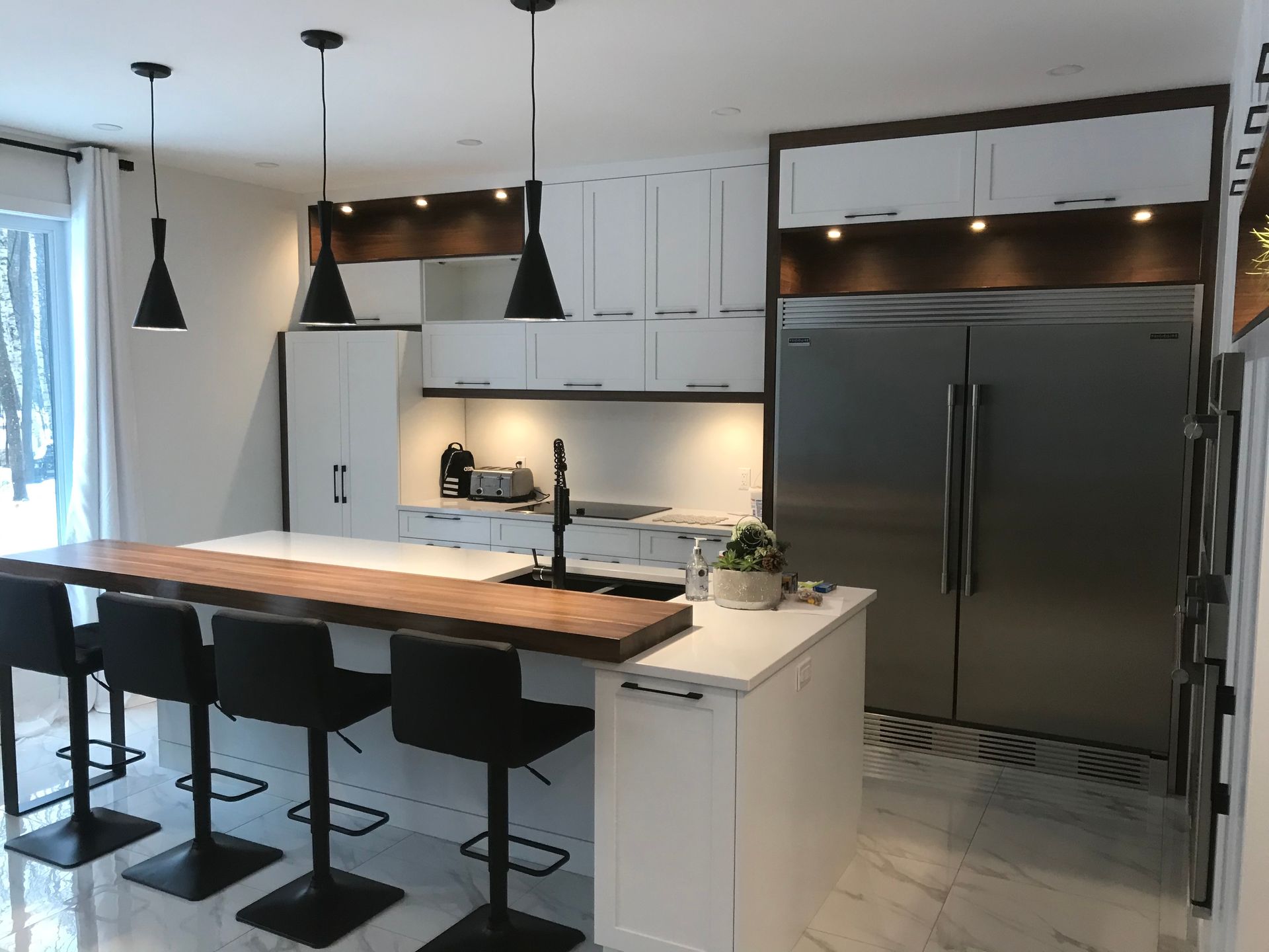 Cuisine blanche moderne avec îlot, réfrigérateur en acier inoxydable, suspensions noires et tabourets de bar.