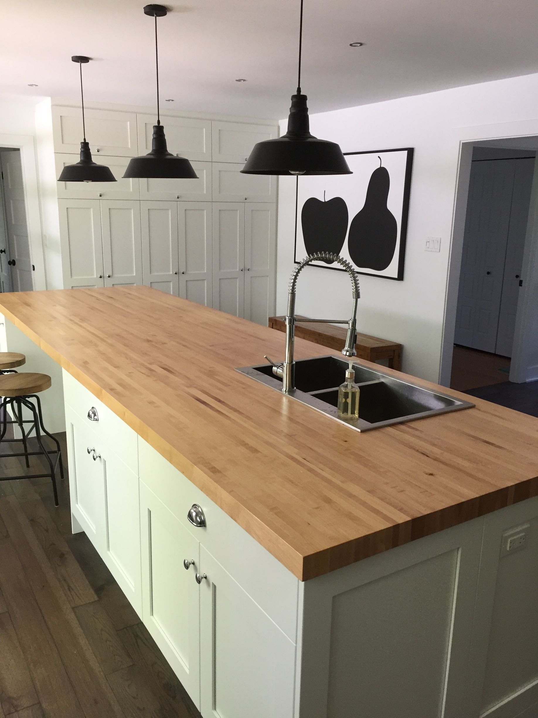 Îlot de cuisine avec comptoir en bois clair, armoires blanches, luminaires suspendus noirs et évier en acier inoxydable.