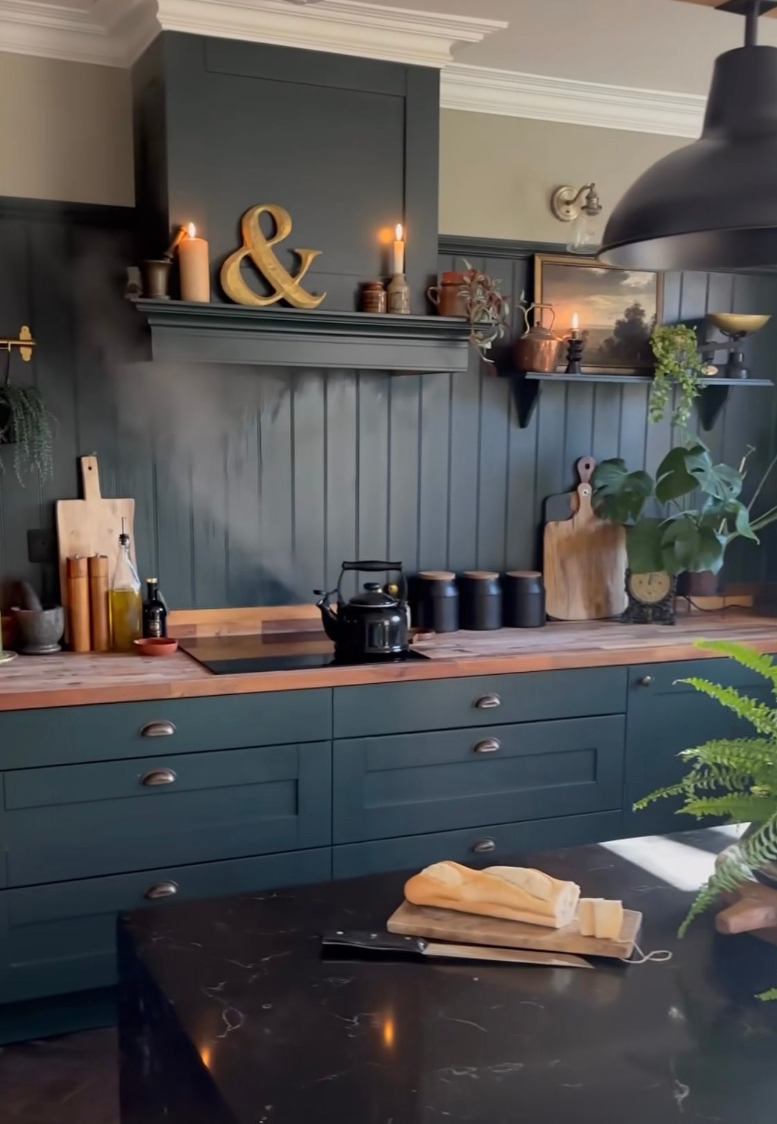 Cuisine bleu sarcelle foncé avec comptoirs en bois, îlot noir, bougies et plantes.