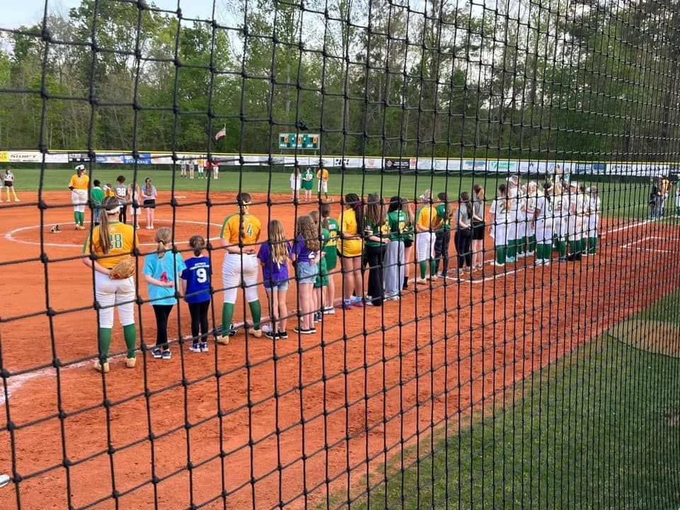 A person wearing a hat and orange shirt, holding a baseball bat on their shoulder on a baseball field.