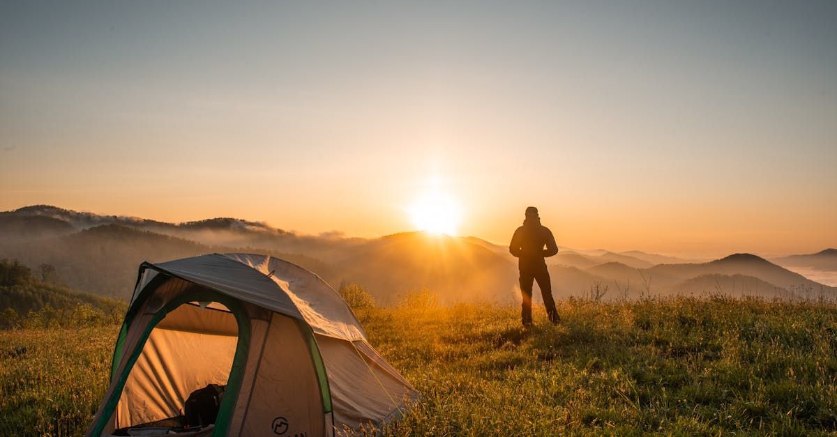 A man is standing in front of a tent on top of a hill at sunset.