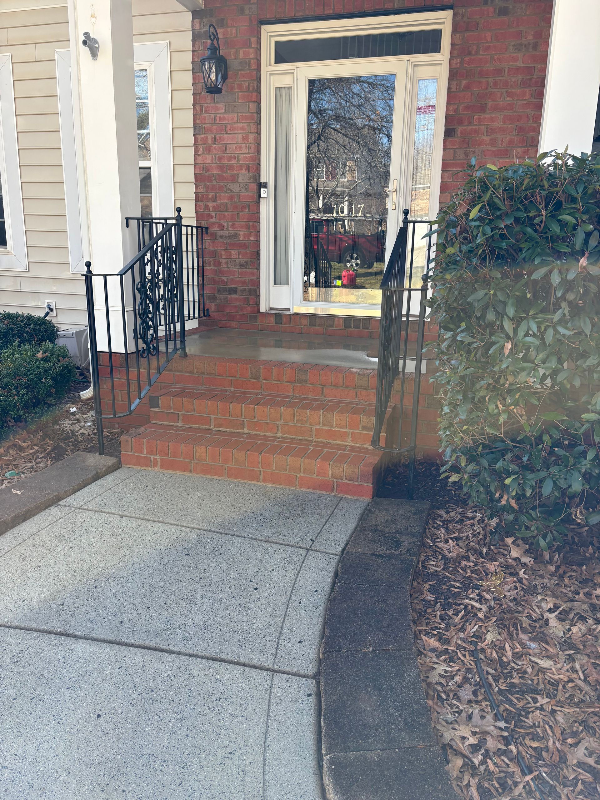 Brick home entrance with steps and railings. Stone walkway and shrubbery are visible.