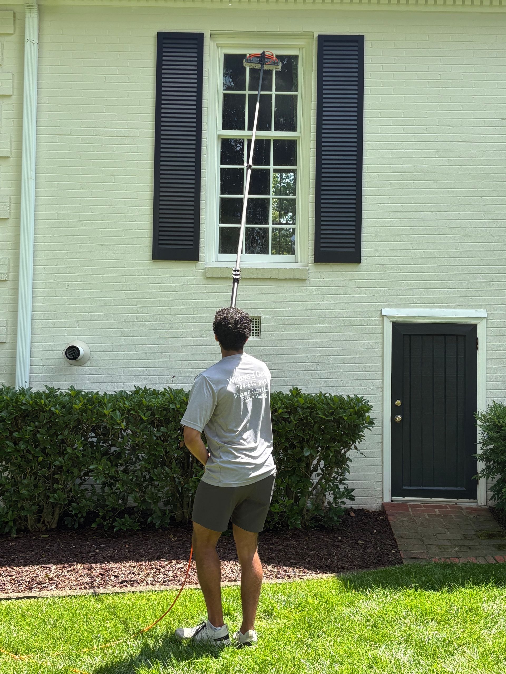 A person washing a window with a long brush outside a white house.