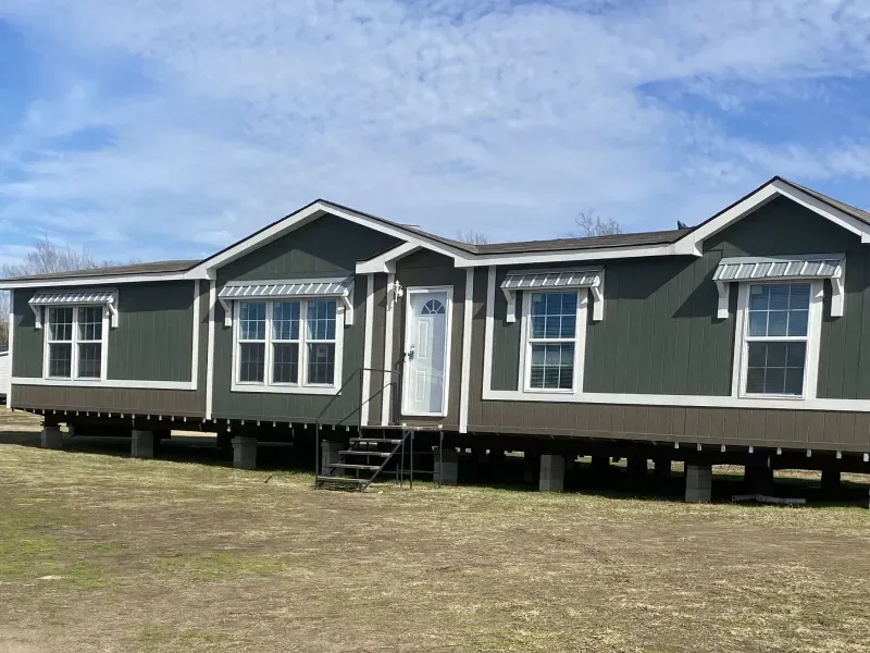 A green and brown mobile home is sitting on stilts in a grassy field.