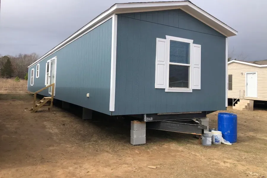 A blue mobile home with white shutters is parked in a dirt field.