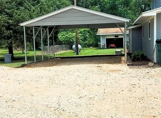 A carport is sitting in the middle of a gravel driveway next to a house.