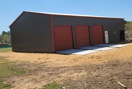 A large metal building with three red garage doors is sitting in the middle of a dirt field.