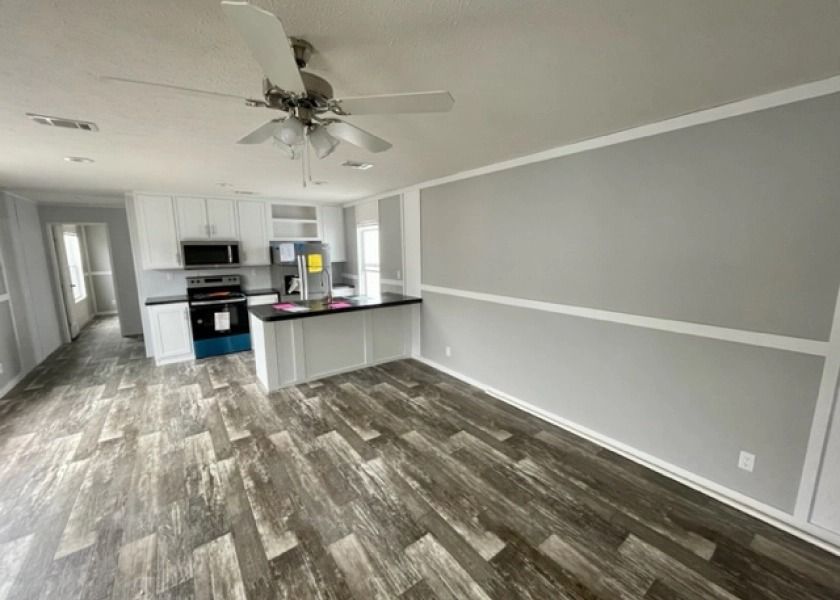 A living room with a ceiling fan and a kitchen in the background.