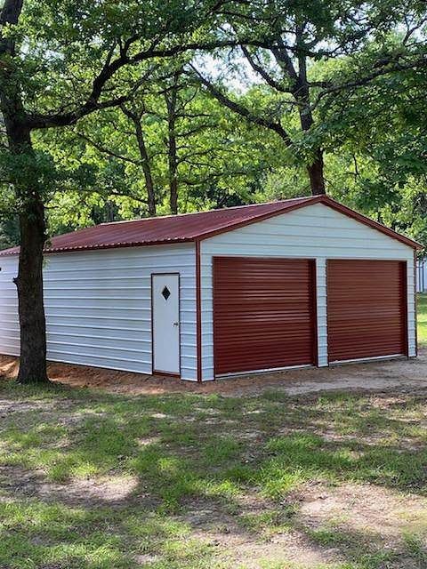 A white garage with red doors is sitting in the middle of a grassy field.